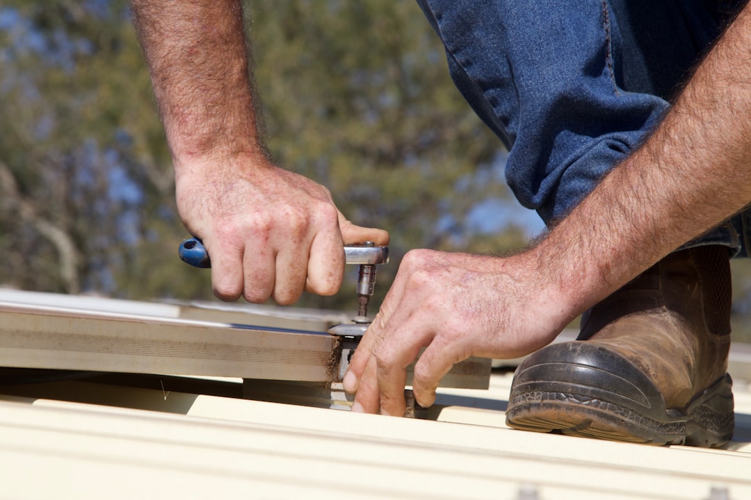 A detailed close-up shot of a construction worker's hands using a nail gun to properly install new architectural asphalt shingles on a home's roof.